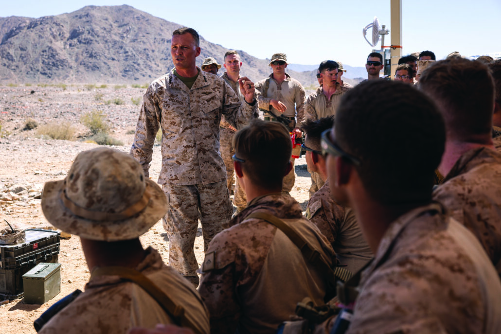 Marine standing in desert environment surrounded by other marines while delivering words.